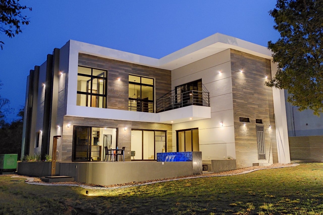 Fachada de vivienda moderna de dos plantas en revestimiento de piedra y madera, iluminada al atardecer, Aqua Village - OGA Constructora